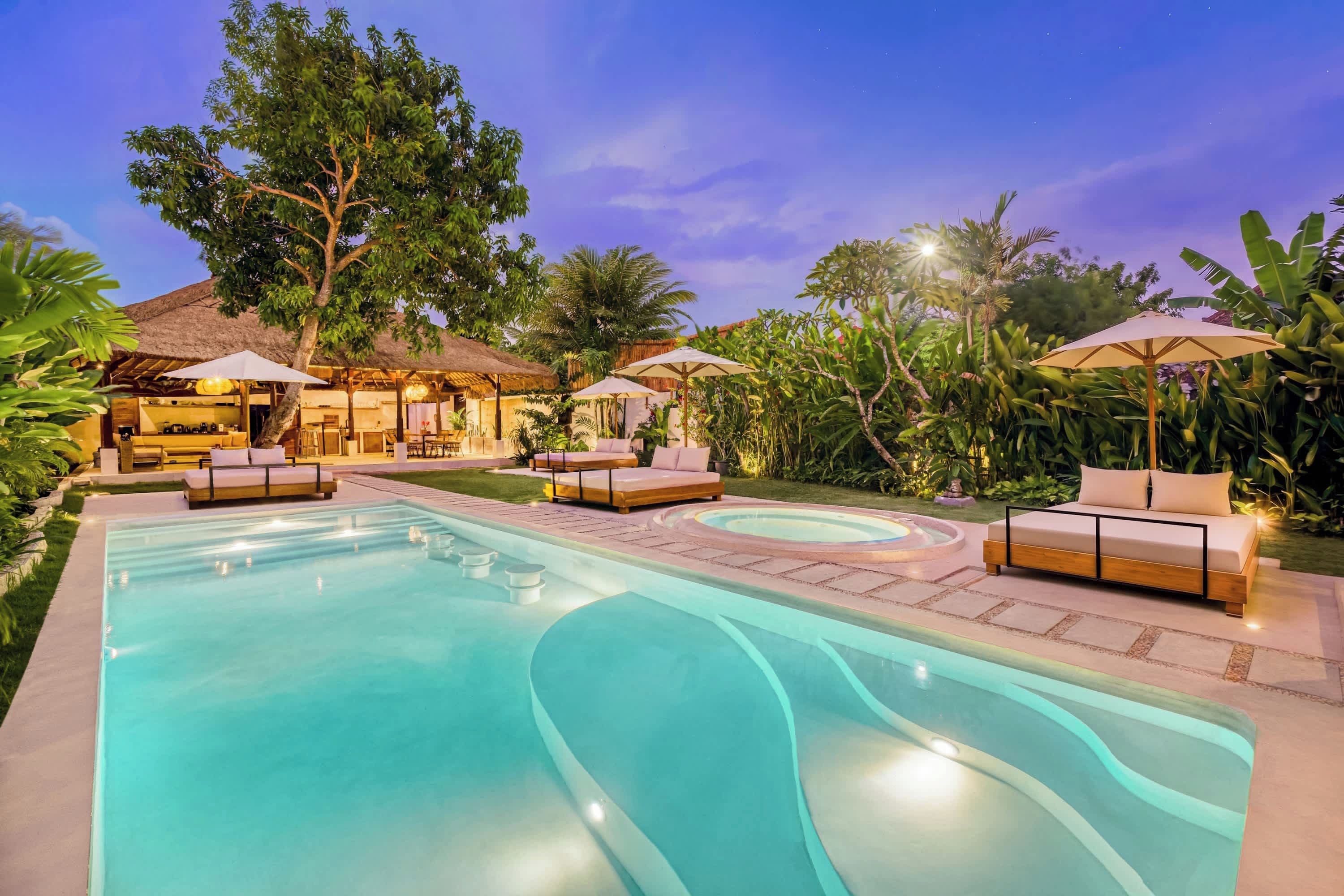 A swimming pool with lounge chairs and umbrellas, surrounded by tropical vegetation and a thatched-roof structure.