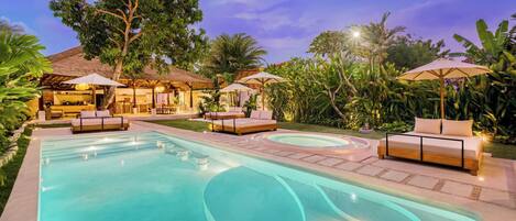 A swimming pool with lounge chairs and umbrellas, surrounded by tropical vegetation and a thatched-roof structure.