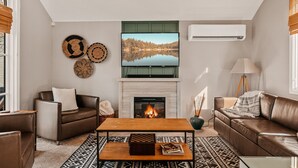 Een woonkamer met een open haard, een televisie erboven en een groot raam met zonwering.