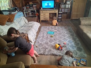 Un salon chaleureux avec un canapé, une table basse et un tapis. Des enfants jouent avec des jouets, et une télévision est allumée en arrière-plan.