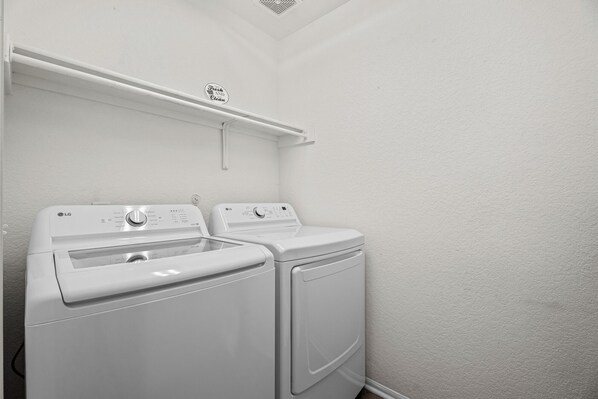 Un área de lavandería con dos lavadoras LG blancas, un estante blanco arriba y una pared blanca con textura.