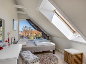 A bedroom with a sloped ceiling, a bed, a desk, and a window overlooking a tree.