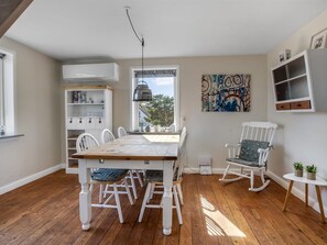 Un coin repas avec une table et des chaises en bois, un fauteuil à bascule blanc, une étagère et un tableau accroché au mur.