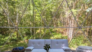 Un espacio exterior moderno y resguardado con un sofá blanco y una mesita pequeña, rodeado de una estructura metálica y vistas a un bosque frondoso.