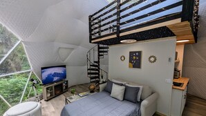 Un dormitorio moderno y funcional con cama, sofá y televisión. Una escalera de caracol conduce a una litera superior.
