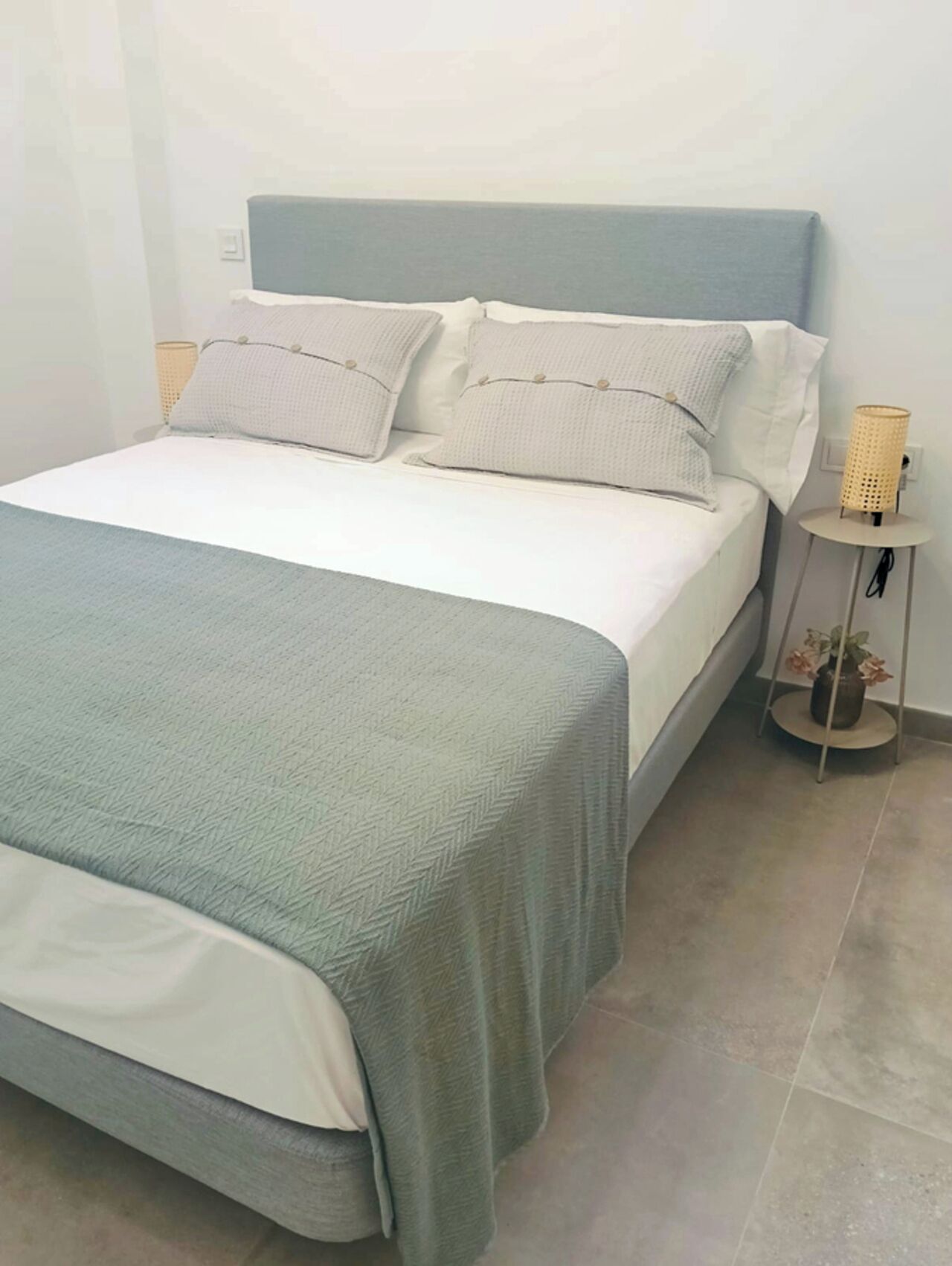 Una cama con cabecera gris y ropa de cama blanca, una mesita redonda con una planta y una lámpara de mimbre al lado de la cama.