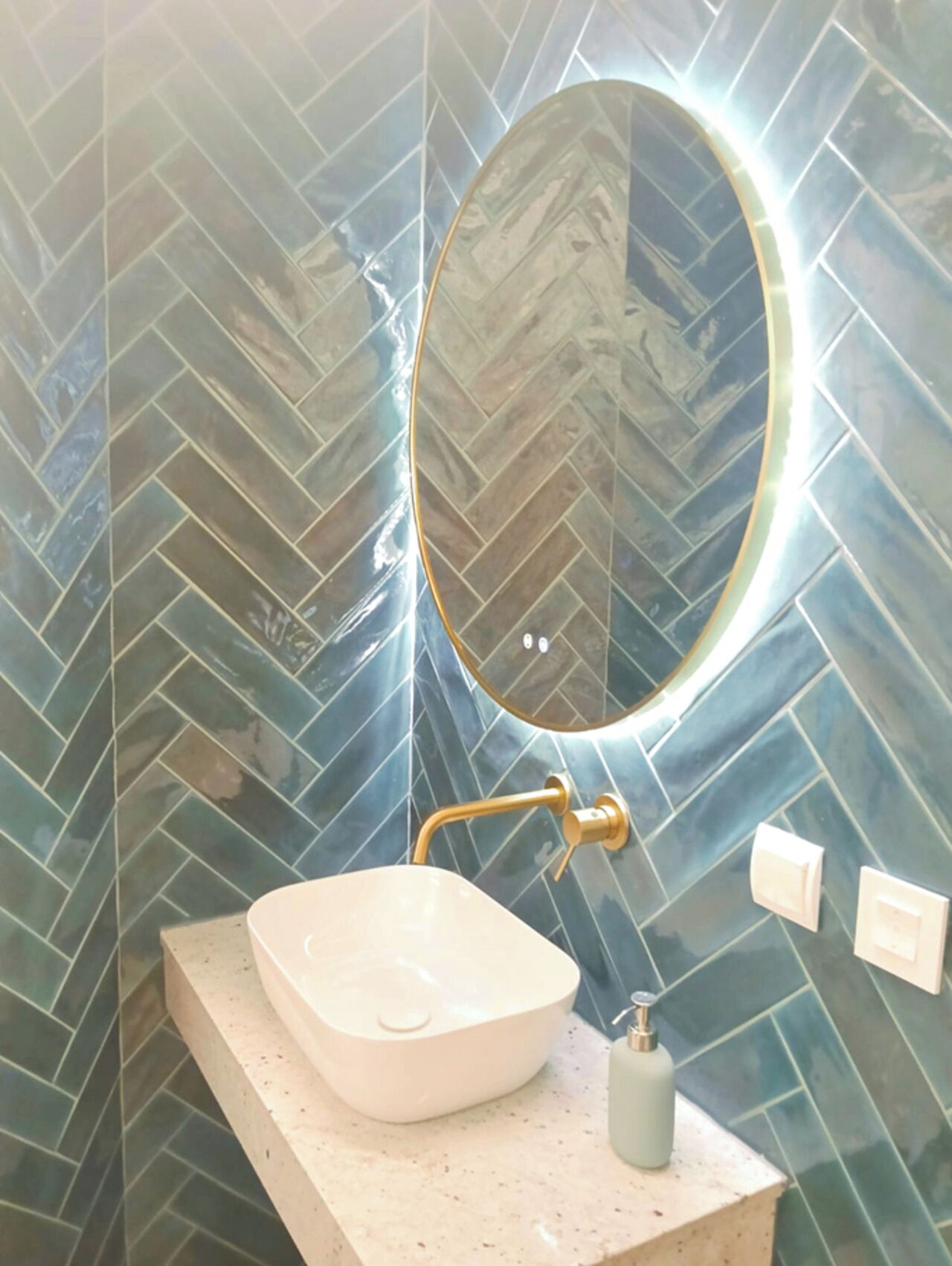 Un baño con detalles de azulejos tipo espiga, un lavamanos blanco de diseño, un grifo dorado, un espejo redondo con luz y un dispensador de jabón.