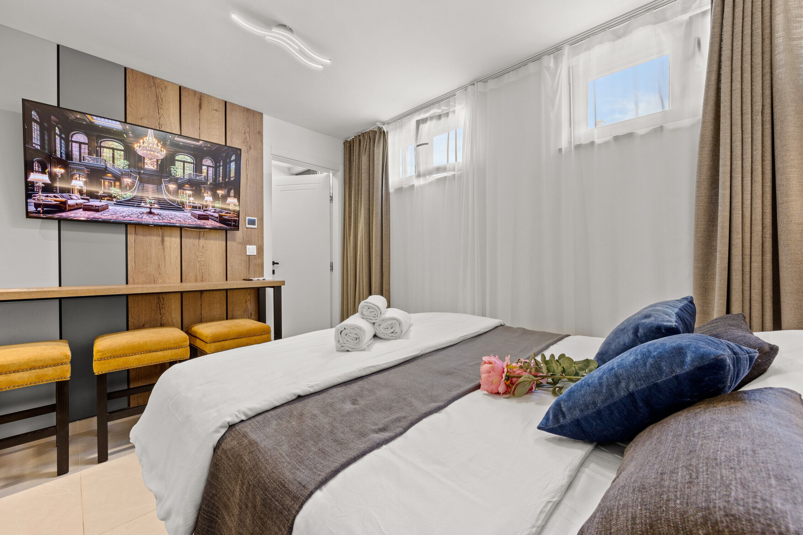 A hotel room with a bed, a TV mounted on the wall, a desk with a chair, and a window with curtains.
