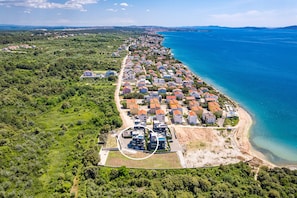 Oversigtsbillede af et kystnære boligområde med huse langs kystlinjen, et klart blå hav og grøn vegetation omkring.