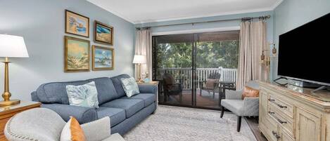 A living room with a sofa, armchair, and a TV on a wooden cabinet. There is a glass door leading to an outdoor area with a patio.