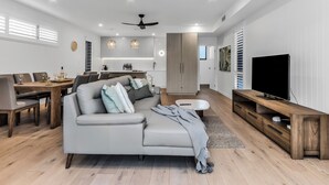 Una sala moderna con un sofá gris, una mesa de centro de madera y un televisor de pantalla plana. Hay un comedor con una mesa y sillas de madera, y una cocina con armarios blancos y lámparas colgantes.