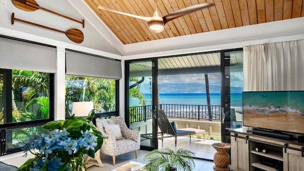 A modern living room with a large TV, comfortable seating, and a balcony with a view of the ocean.