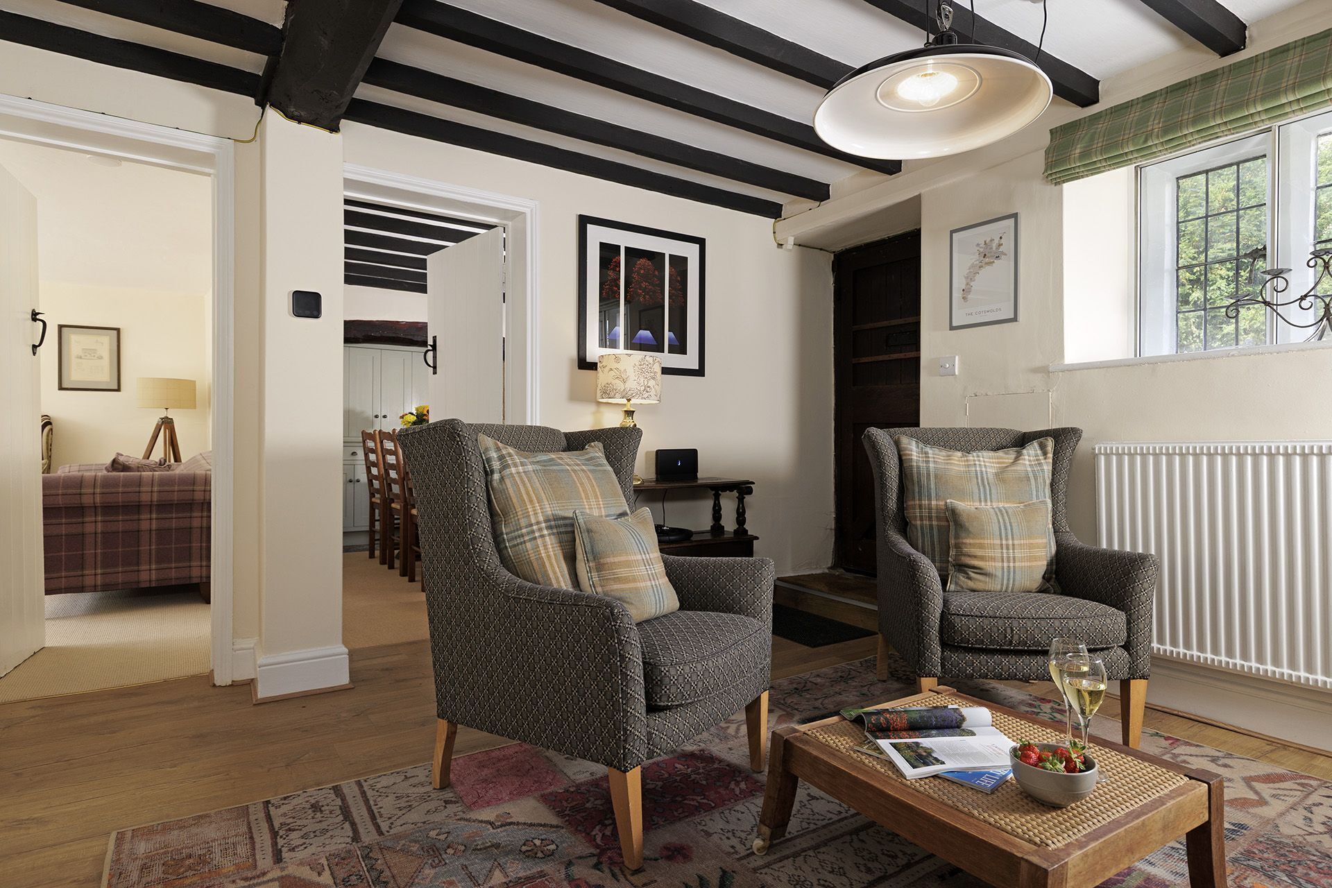 A cozy living room with two patterned armchairs, a coffee table with a book and a glass of wine, and a view of a dining area through an open door.