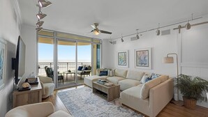Ein geräumiges Wohnzimmer mit großem Fenster, Deckenventilator und Balkon mit Gartenmöbeln.