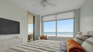 Ein Schlafzimmer mit einem großen Bett, einem Deckenventilator, einem Fernseher und einem Fenster mit Blick auf den Strand.