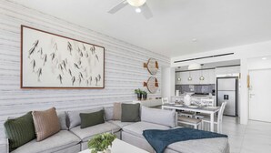 A modern living room with a grey sectional sofa, a white coffee table, and a kitchen in the background. - Marea | Medano Beach Cabo Luxury Condo Rental (Cabo San Lucas)