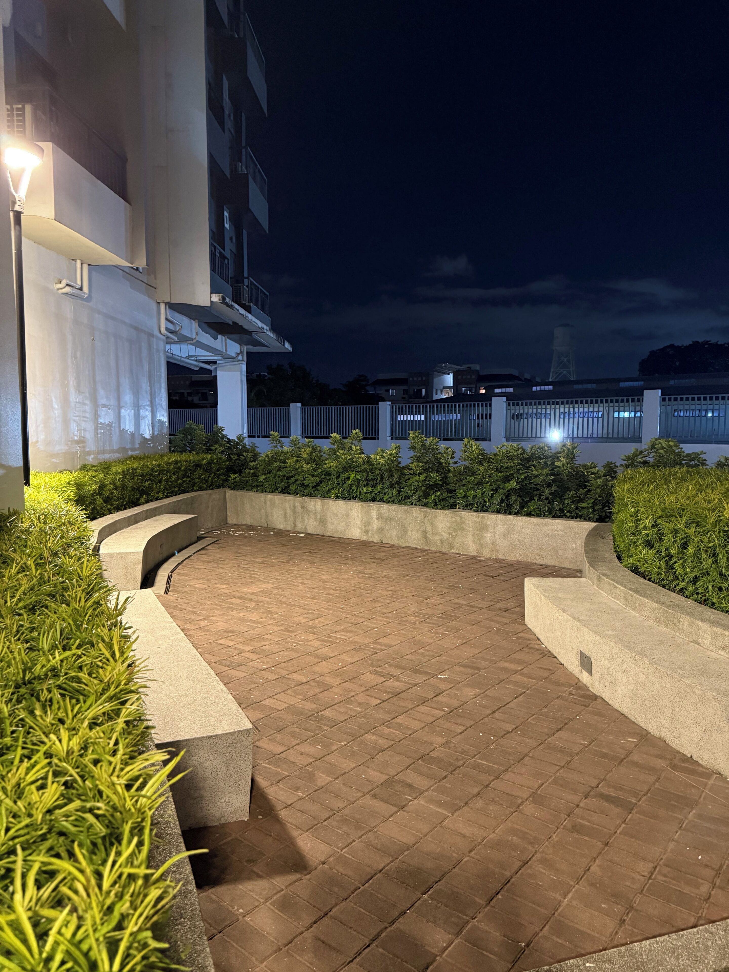 A nighttime outdoor area with a paved walkway, low-rise buildings, and a distant view of a body of water.
