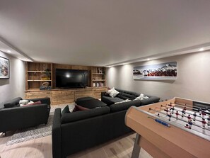 Uma sala de estar moderna com uma televisão de tela grande, um conjunto de sofás, um móvel de entretenimento de madeira e uma mesa de pebolim.