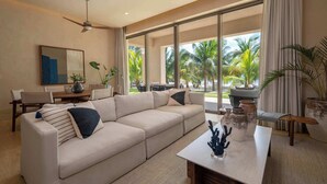 A spacious living room with a beige sectional sofa, a dining table, and a ceiling fan. Large windows offer a view of palm trees outside.