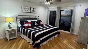 Un dormitorio con cama, ventilador de techo, mesita de noche, lámpara, ventana con cortinas y vistas al exterior.