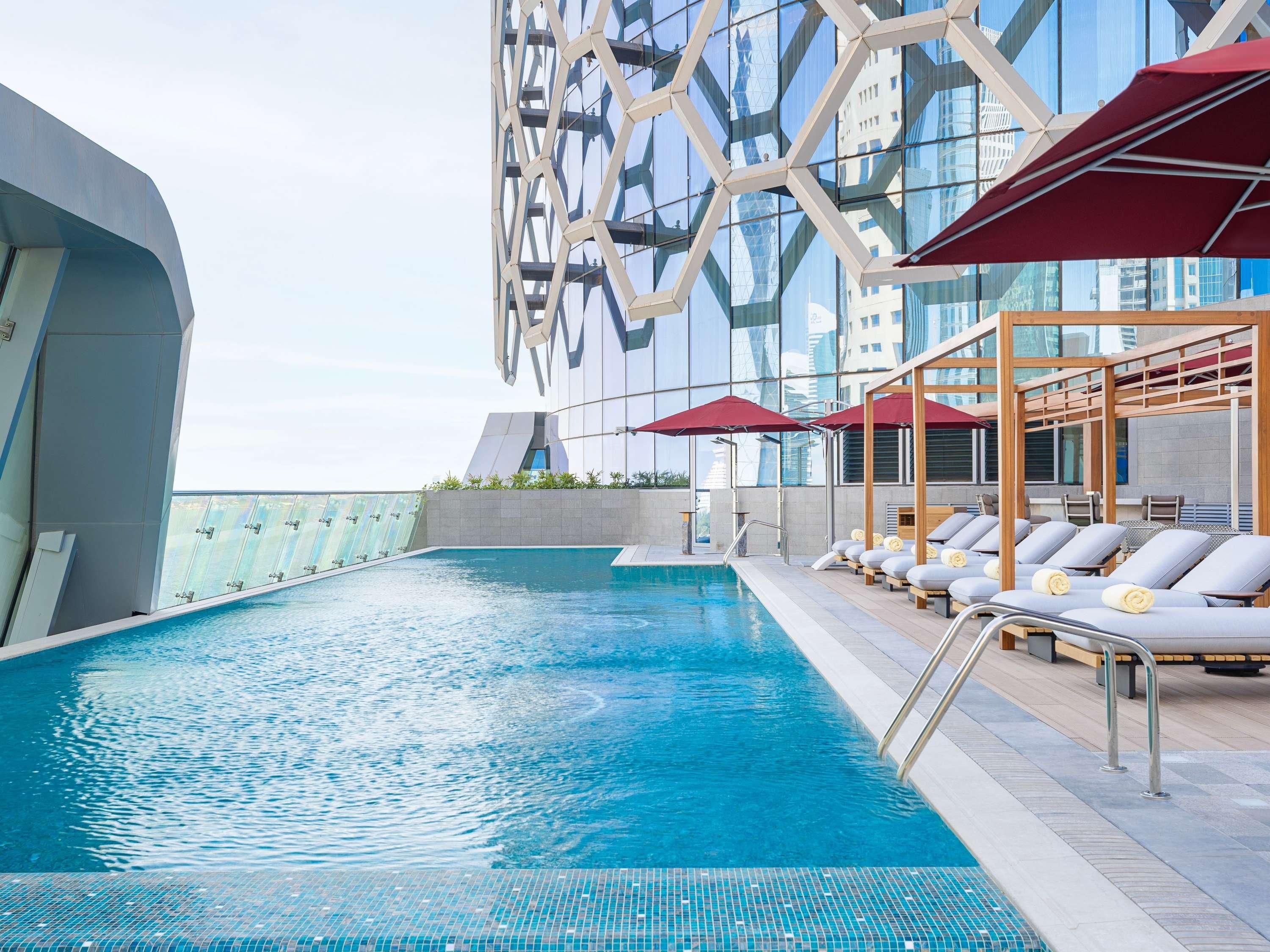A rooftop swimming pool with lounge chairs and umbrellas, surrounded by a modern building with reflective glass windows.