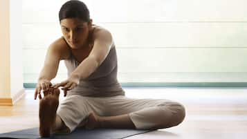 Uma pessoa sentada num tapete de yoga numa divisão com soalho de madeira e uma janela grande.