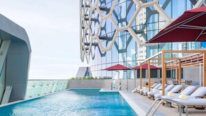 Une piscine sur le toit, agrémentée de chaises longues et de parasols, entourée d’un bâtiment moderne aux fenêtres en verre réfléchissant.