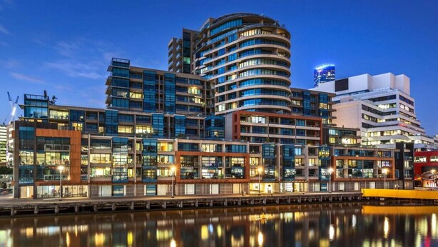 A modern urban building complex with multiple high-rise structures, a waterfront promenade, and a reflection in the water.
