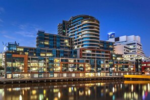 Un moderno complejo de edificios urbanos con varias torres altas, un paseo marítimo y un reflejo en el agua.