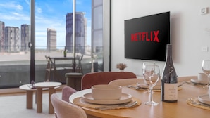 A modern dining area with a table set for two, a wine bottle, and a television displaying the Netflix logo.