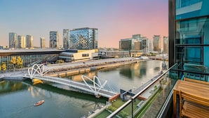 A modern building with a glass facade overlooking a river, a pedestrian bridge, and a view of the city skyline.