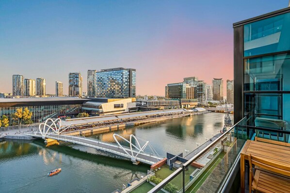 Un edificio moderno con fachada de vidrio que da a un río, un puente peatonal y vistas del horizonte de la ciudad.