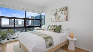 A modern bedroom with a large bed, a bedside table with a lamp, and a city view through floor-to-ceiling windows.