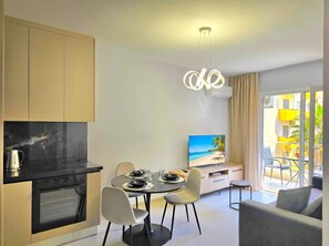 A modern kitchen with a built-in oven, a dining area with a table and chairs, a TV on a stand, and a balcony with a view of buildings and palm trees.