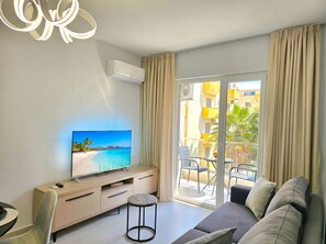 A modern living room with a flat-screen TV, a sofa, a small round table, and a balcony with outdoor furniture.