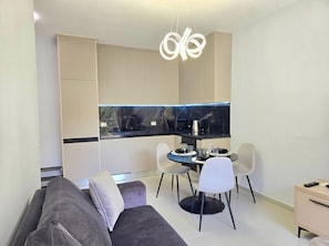 Una sala de estar moderna con un sofá, una mesa de comedor con sillas, una zona de cocina con encimera negra y una lámpara colgante.