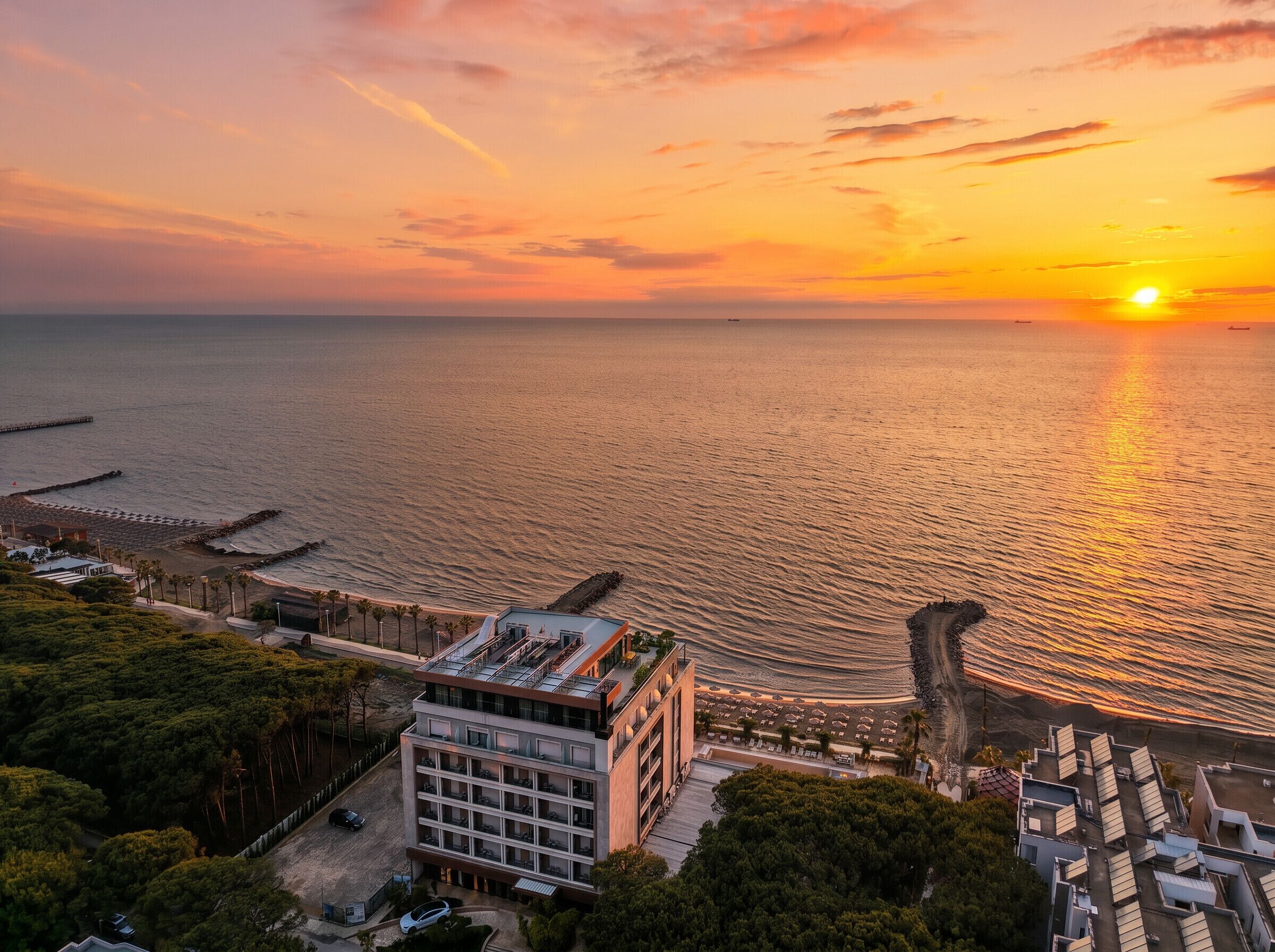 Um hotel à beira-mar com vista para o pôr do sol sobre o oceano, uma área de estacionamento e um edifício com vários andares.