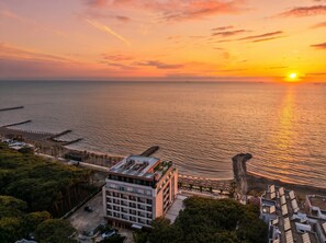 A coastal hotel with a view of the sunset over the sea, a parking area, and a building with multiple floors.