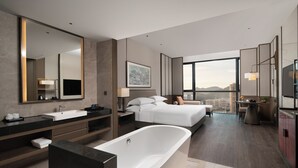 A modern hotel room with a large bathtub, a double bed, a sitting area with a table and chairs, and a large window offering a city view. - Juntels Panan Jinhua (Jinhua)