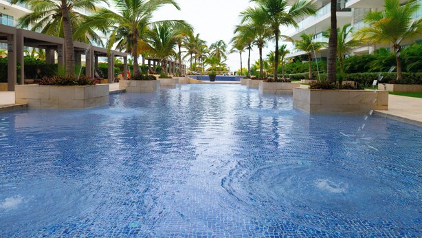 Una amplia piscina interior con suelo de baldosas, rodeada de palmeras y edificios modernos.