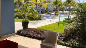 Vestíbulo moderno de hotel con un amplio ventanal que ofrece vistas a una piscina, palmeras y un edificio al fondo.