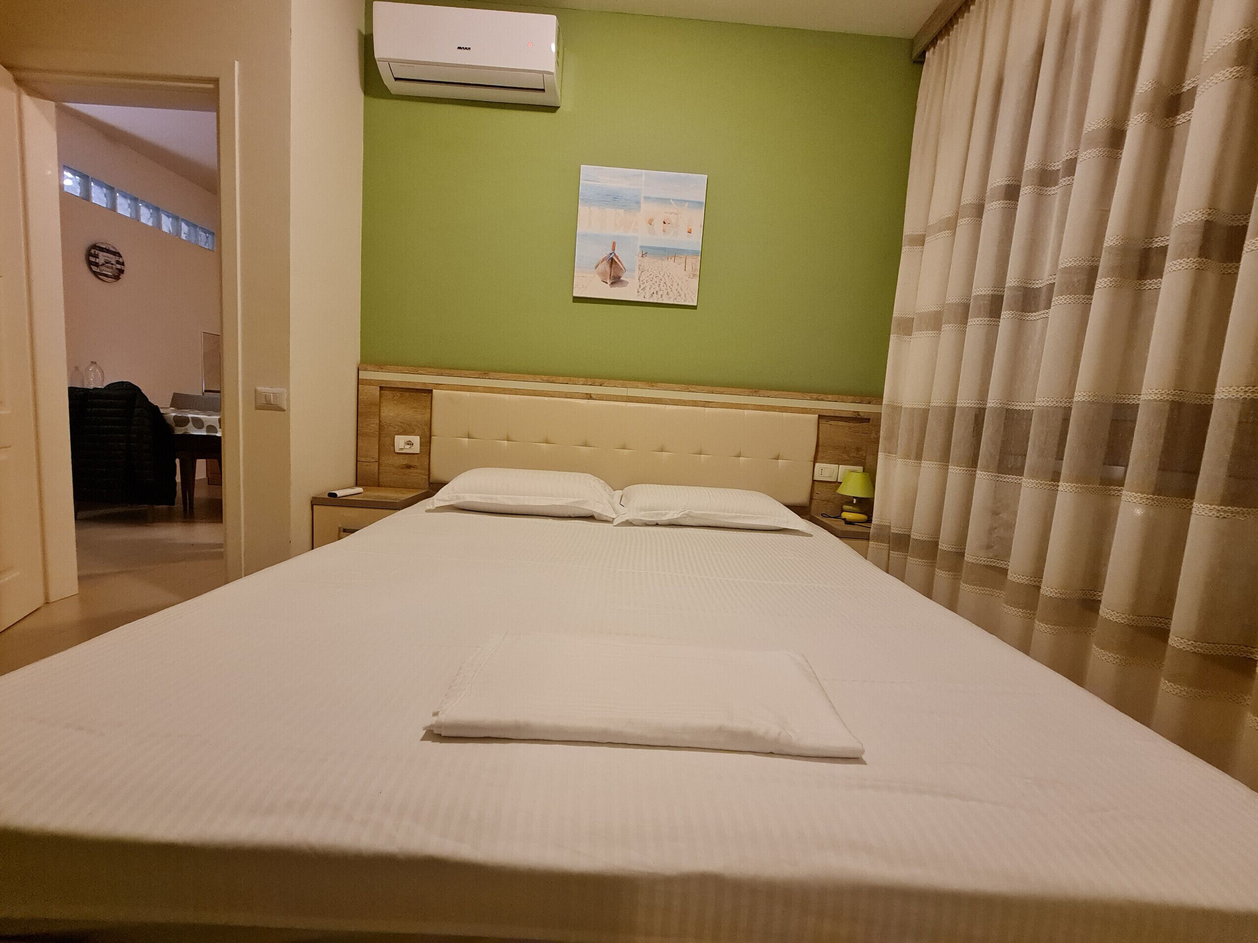 Un lit bien fait dans une chambre d’hôtel, avec un mur vert, un tableau et une fenêtre avec des stores.