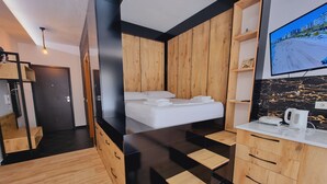 A modern hotel room with a bed, wooden wardrobe, and a kitchen area with a TV. - Teo Deluxe Studios - Family Triple  Studio (Vlorë)