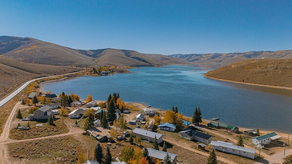 Un poblado junto al lago con casas y cabañas, rodeado de colinas y un cielo despejado.