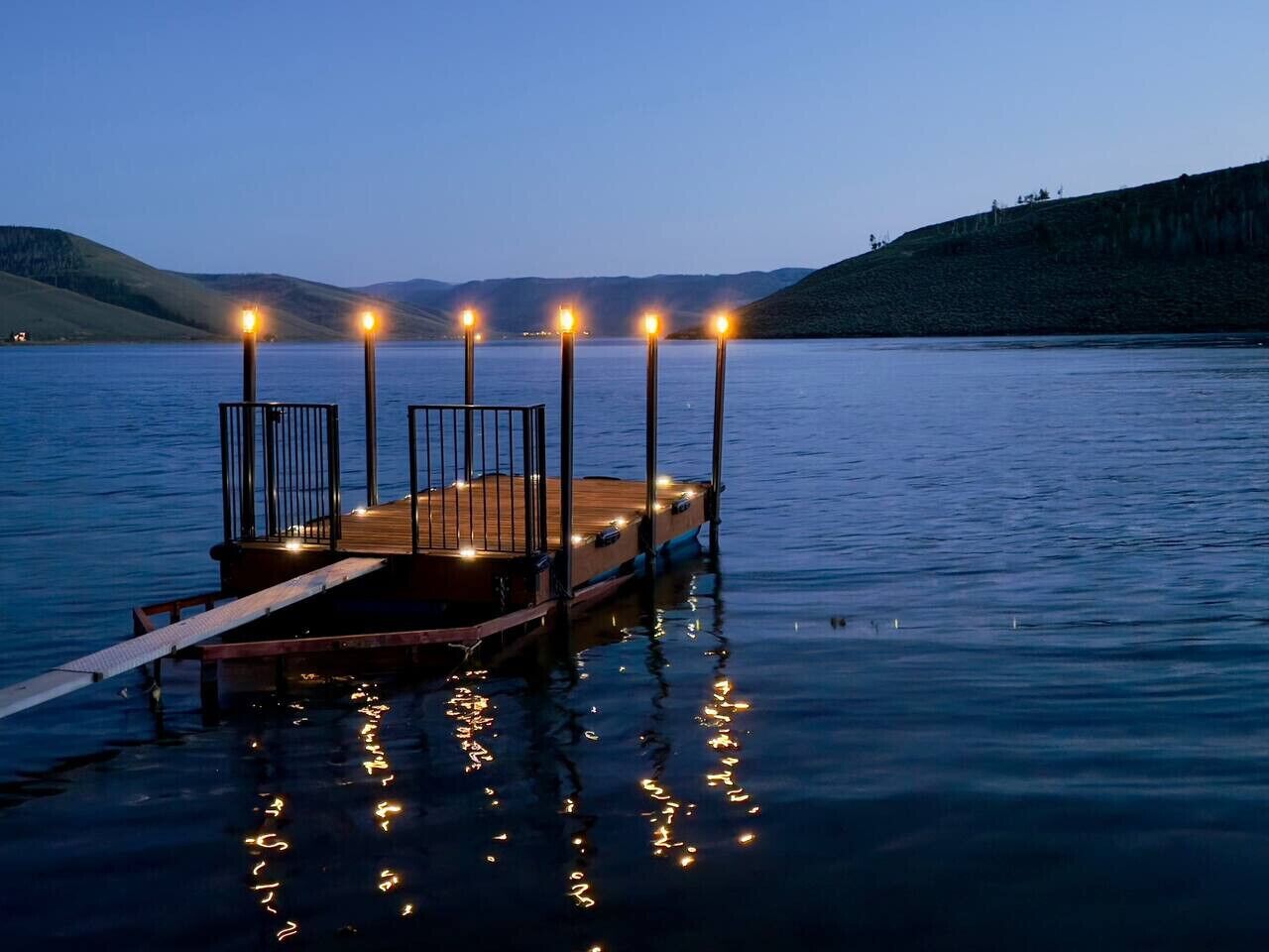 Ein Steg, der sich in ruhiges Wasser erstreckt, mit einer Holzplattform und Geländern, umgeben von Hügeln.
