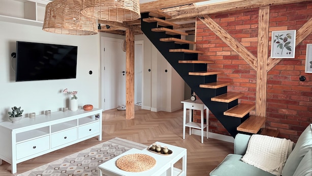 A modern living room with a white sofa, a coffee table, and a TV on the wall. There's a staircase with a wooden railing and a brick wall.