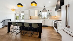 A modern kitchen with white cabinets, a large island with bar stools, a stainless steel refrigerator, and pendant lighting. - Thoughtfully Designed Luxury Group Retreat (Ephraim)
