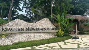 A sign for Mactan Newtown Beach with a stone wall backdrop, surrounded by lush greenery and a thatched structure.