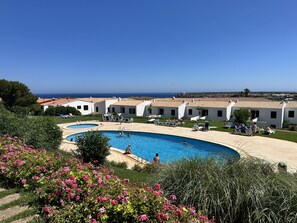 Ein Poolbereich mit zwei Schwimmbecken, umgeben von weißen Gebäuden mit roten Ziegeldächern und Blick auf das Meer im Hintergrund.