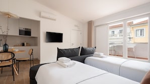 A hotel room with two beds, a kitchenette, a TV, and a balcony with a view of buildings.